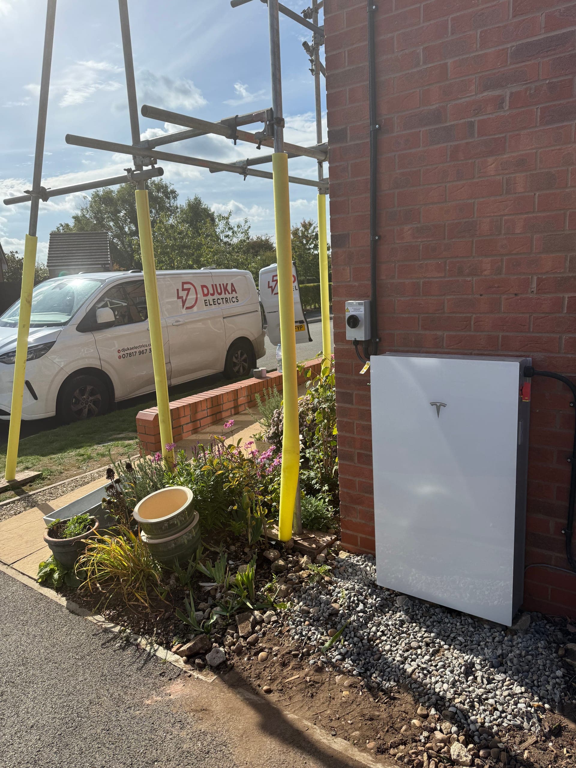 David Clarke's Tesla Powerwall installed on the side of his brick home with the Djuka Electrics van and scaffolding visible behind